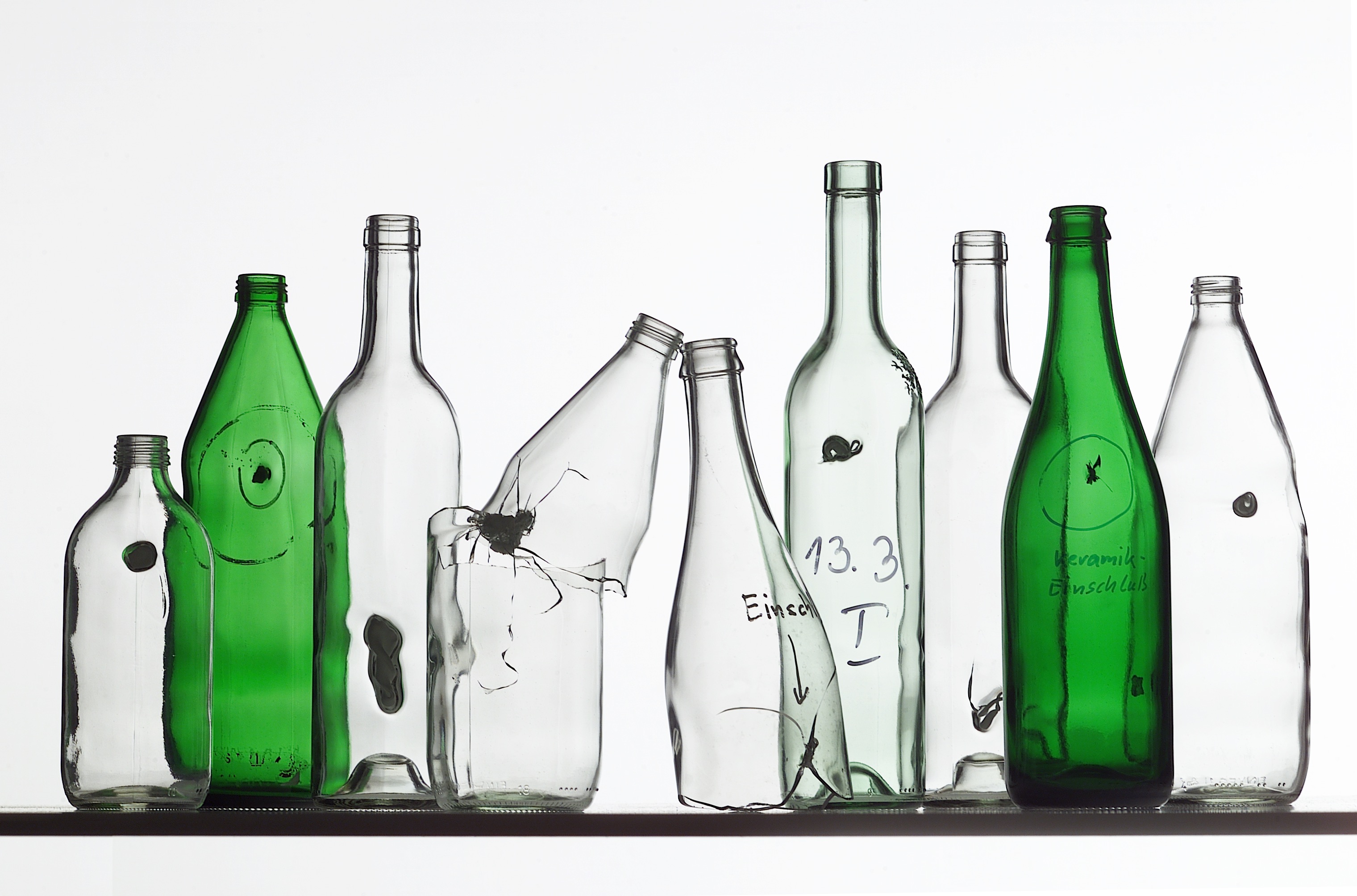 Mehrere durchsichtige und zwei grüne Flaschen, die alle fehlerhaft sind, da sie sogenannte Einschlüsse aus anderen Materialien enthalten.
