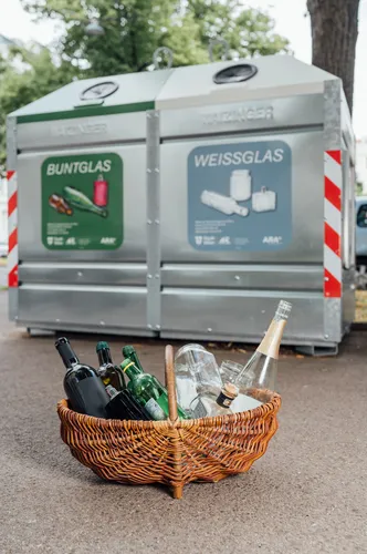 Korb mit leeren Glasverpackungen vor Buntglas- und Weißglas-Containern.